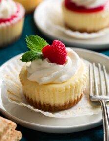 Mini cheesecake shown on a dessert plate unwrapped from cupcake liner. It is topped with whipped cream, fresh raspberry and mint.