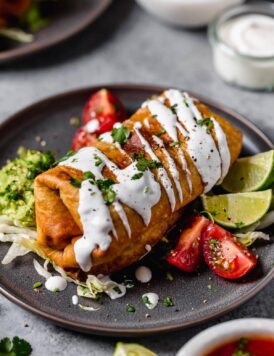 Chicken Chimichanga on a plate. It's drizzled with sour cream, garnished with cilantro and served with tomatoes and guacamole on the side.