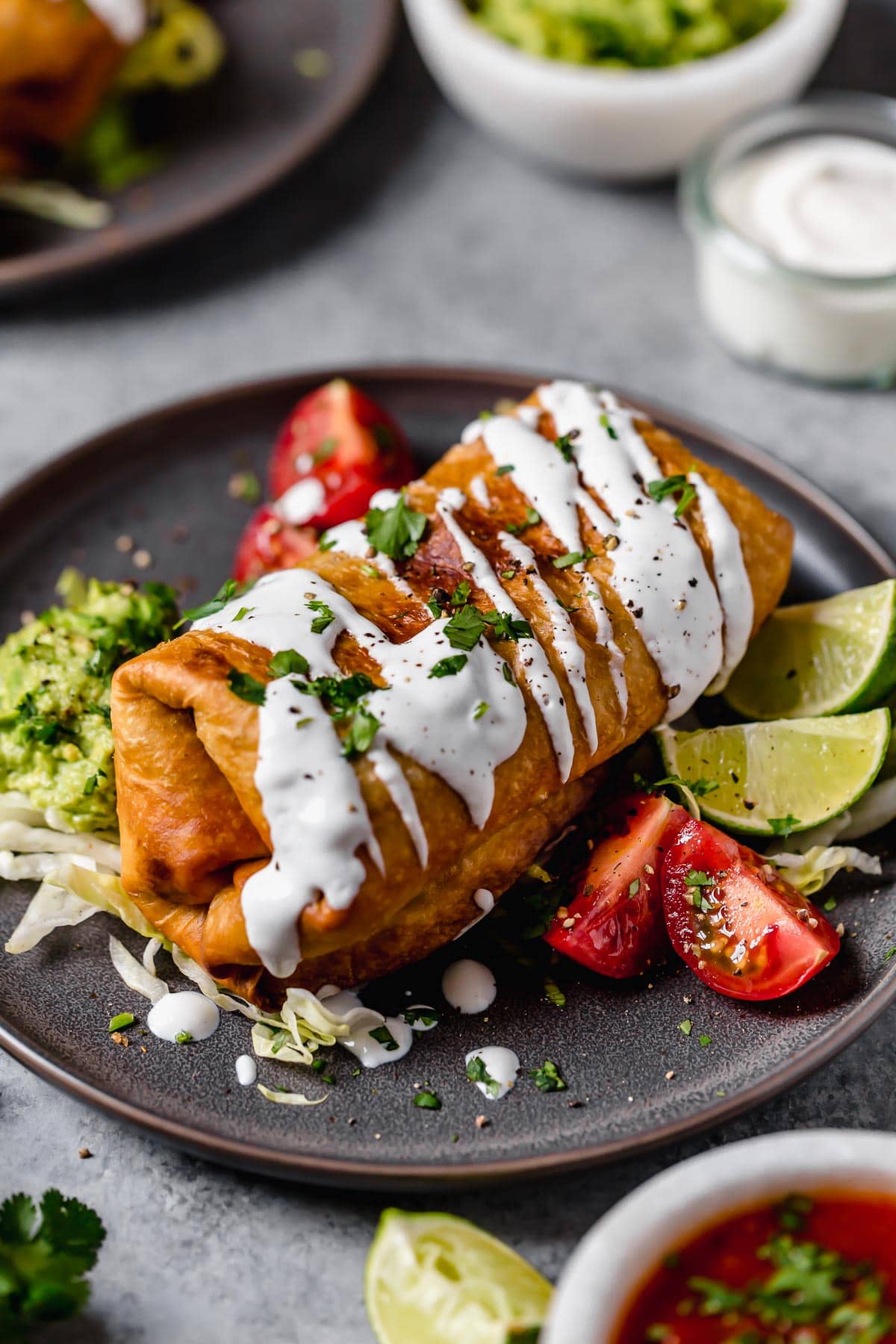 Chicken Chimichanga on a plate. It's drizzled with sour cream, garnished with cilantro and served with tomatoes and guacamole on the side.