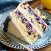 Slice of lemon blueberry cake with cream cheese frosting on a blue plate set over a wooden platter.