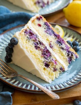 Slice of lemon blueberry cake with cream cheese frosting on a blue plate set over a wooden platter.