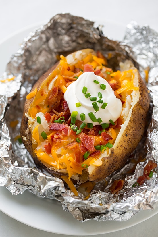 Baked potato made in the slow cooker. Its cut open and topped with cheddar, bacon, sour cream and chives. Shown sitting on a white serving plate.