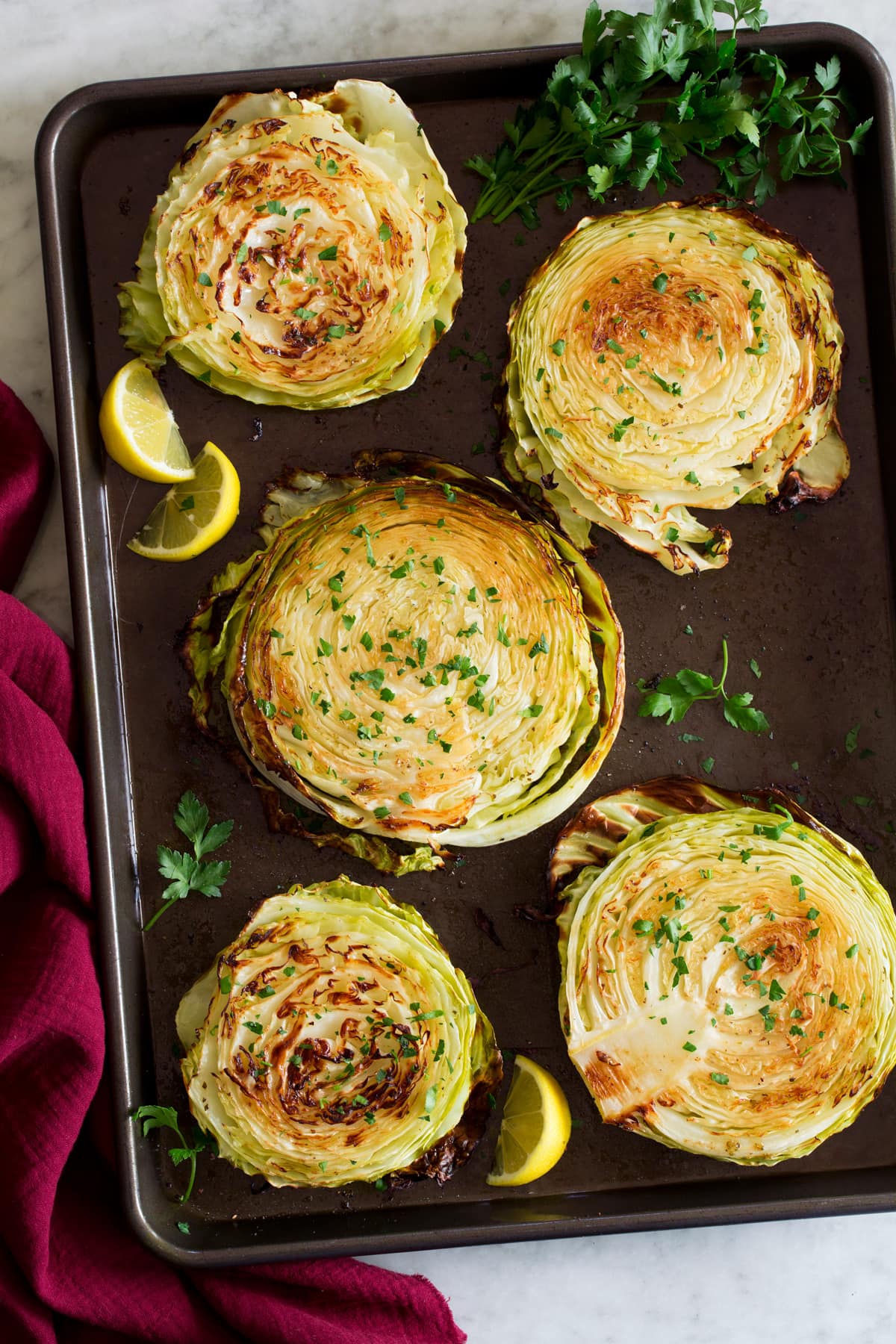 Photo: Shows roasted cabbage slices from overhead on a dark baking sheet. Parsley is shown to the side and a red cloth.