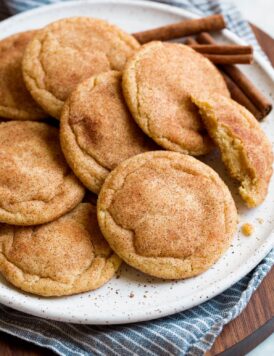 Image of snickerdoodles cookies shown from the side layered together on a white plate with cinnamon sticks in the background. Plate is resting on a striped blue cloth and wooden platter.