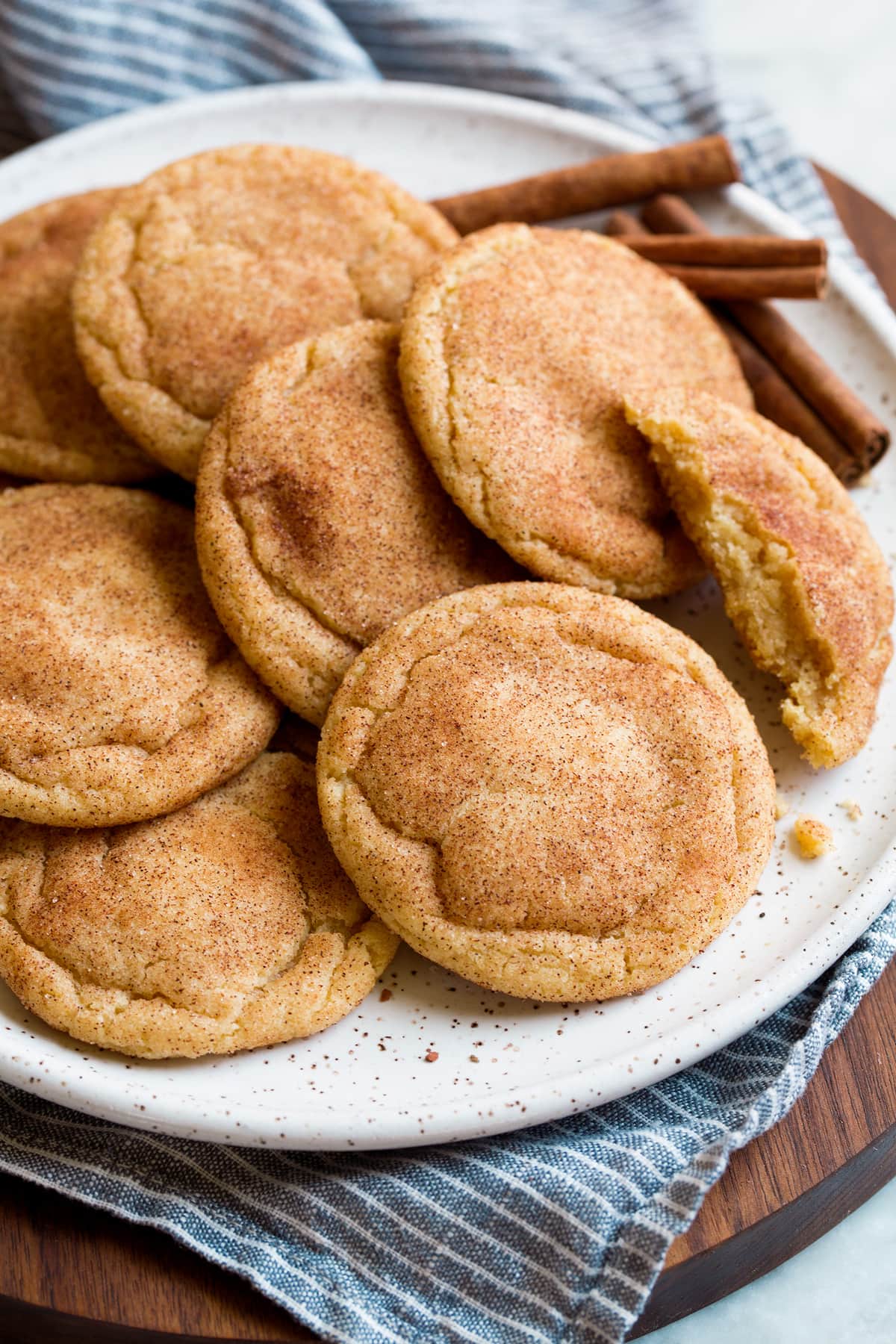 Image of snickerdoodles cookies shown from the side layered together on a white plate with cinnamon sticks in the background. Plate is resting on a striped blue cloth and wooden platter.