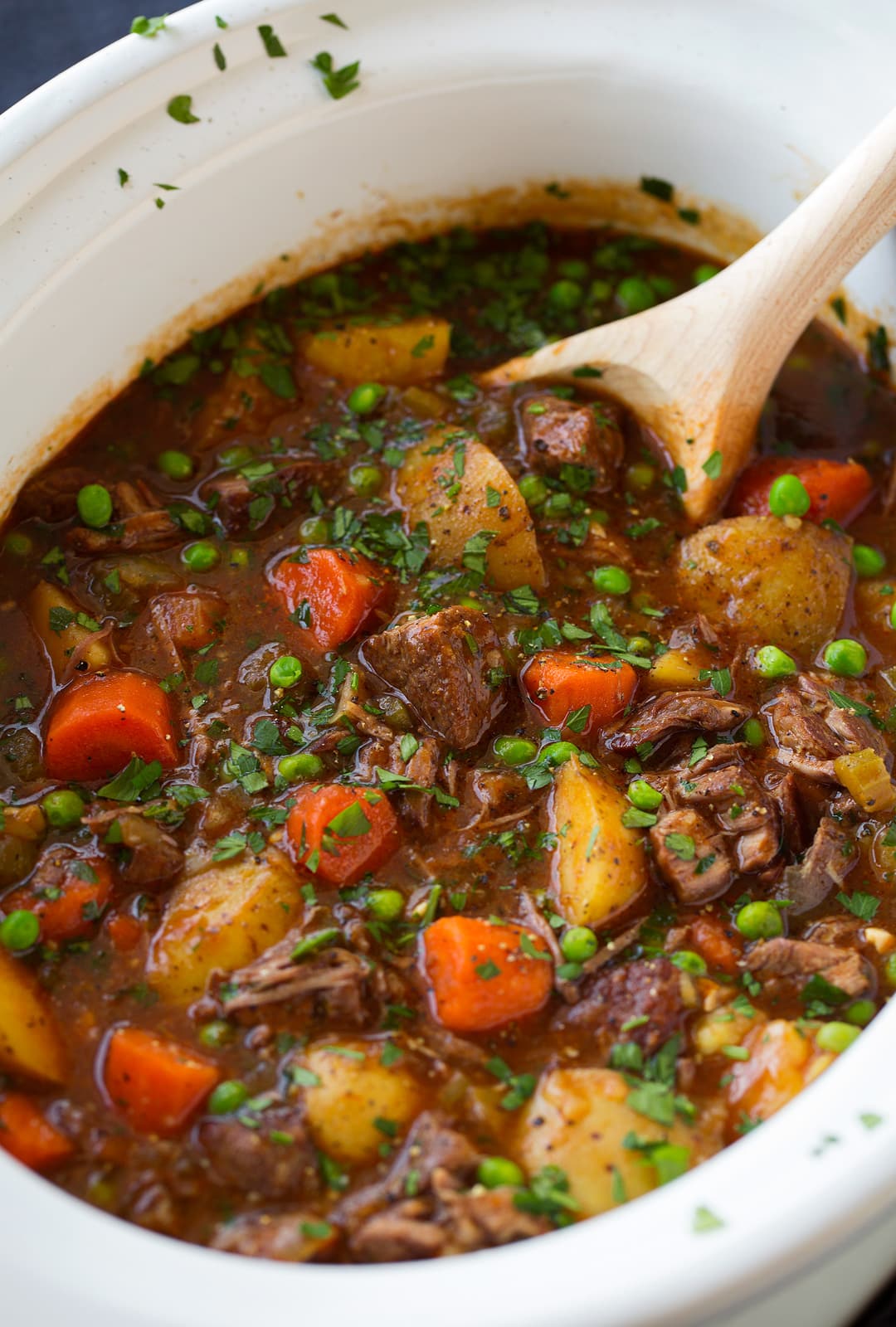 Beef Stew shown here in a white slow cooker with wooden spoon for ladling.