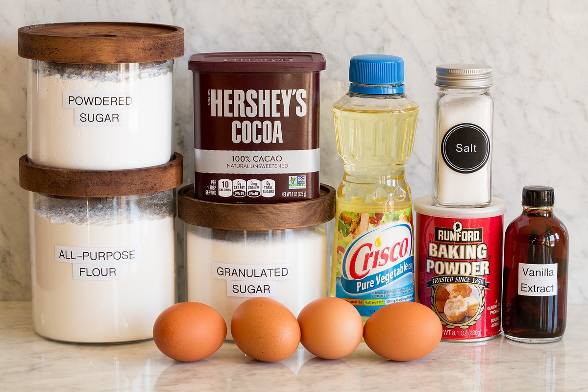 Image of ingredients used to make chocolate crinkle cookies. Includes flour, granulated sugar, powdered sugar, eggs, hershey's cocoa, vegetable oil, baking powder, salt and vanilla extract.