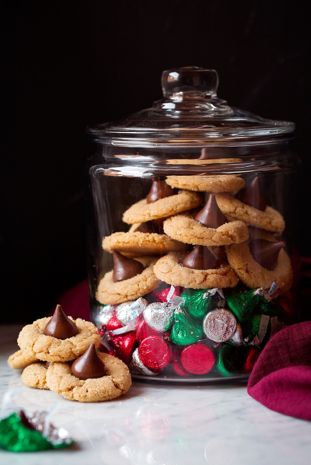 Peanut Butter Blossoms shown here in a clear storage jar over a pile of Hershey's Kisses. Container is set on a marble surface with a black background.