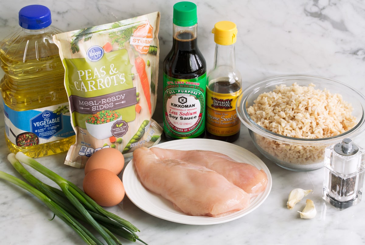 Ingredients needed for chicken fried rice shown here including chicken breasts, brown rice, soy sauce, sesame oil, garlic, eggs, green onions, vegetable oil, frozen peas and carrots.