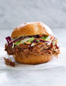 Pulled Pork shown here in a bun with coleslaw sitting on parchment paper on a marble surface