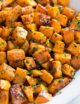 Roasted butternut squash with garlic and fresh herbs in a white ceramic serving bowl set over a white marble surface.