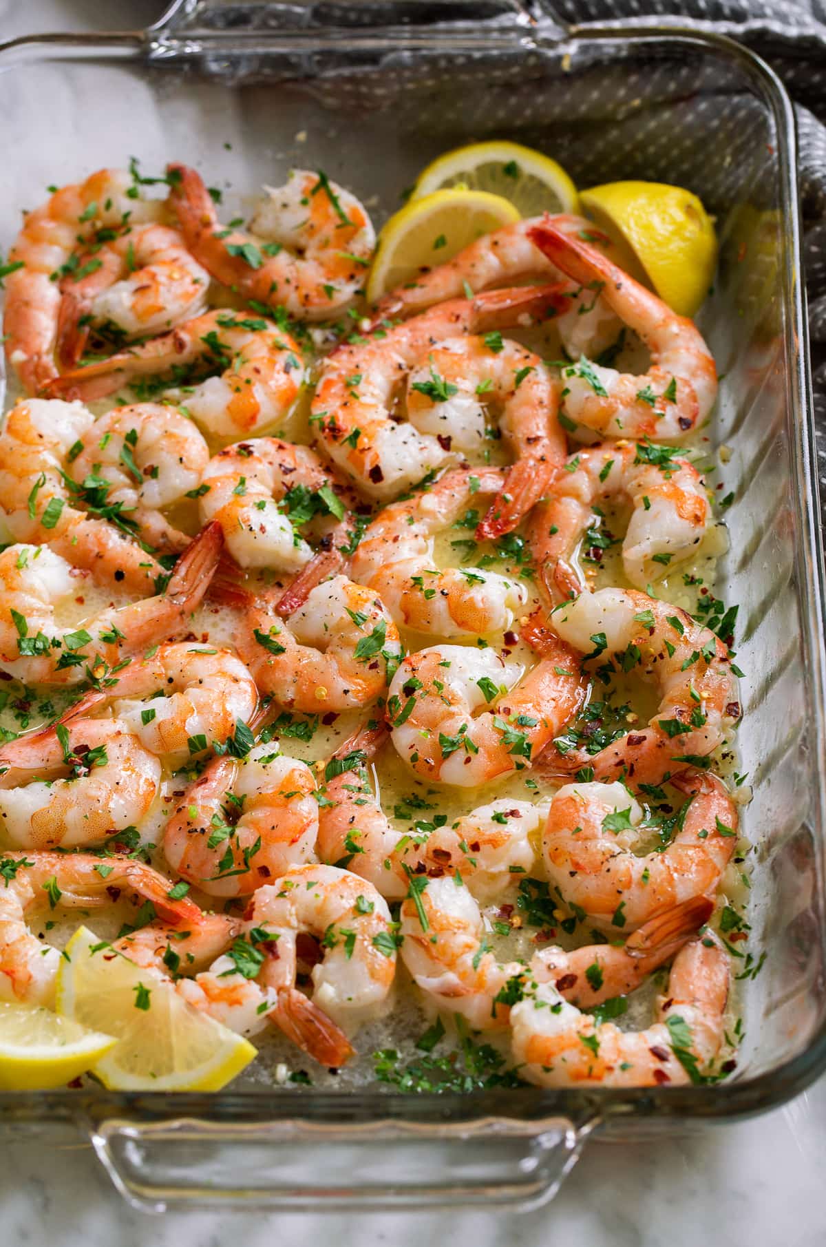 Glass baking dish filled with bake shrimp and garlic butter sauce, garnished with parsley and lemon wedges.