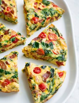 Six frittata wedges shown on a white serving platter set over a marble surface. Frittata is filled with bacon, spinach, tomatoes and swiss.