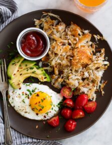 Hash browns on brown serving plate with sliced tomatoes, sliced avocado, fried egg and a side of ketchup.