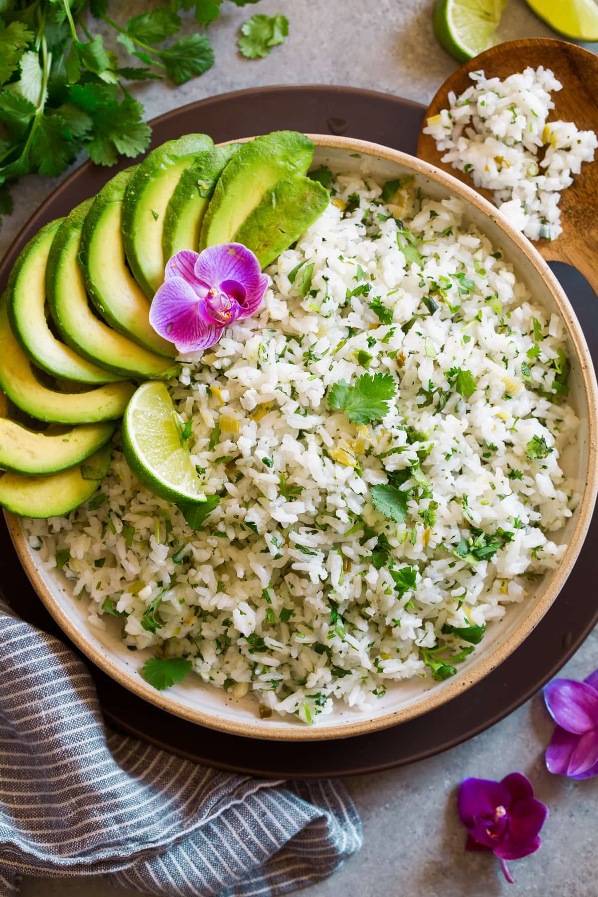 Cilantro Lime Rice in a cream ceramic serving bowl set over a brown matte plate. Rice is garnished with cilantro, lime, avocado and orchid flower.