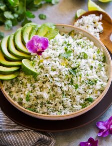 Serving bowl full of cilantro lime rice garnished with avocado slices, cilantro leaves, orchid flower and lime.