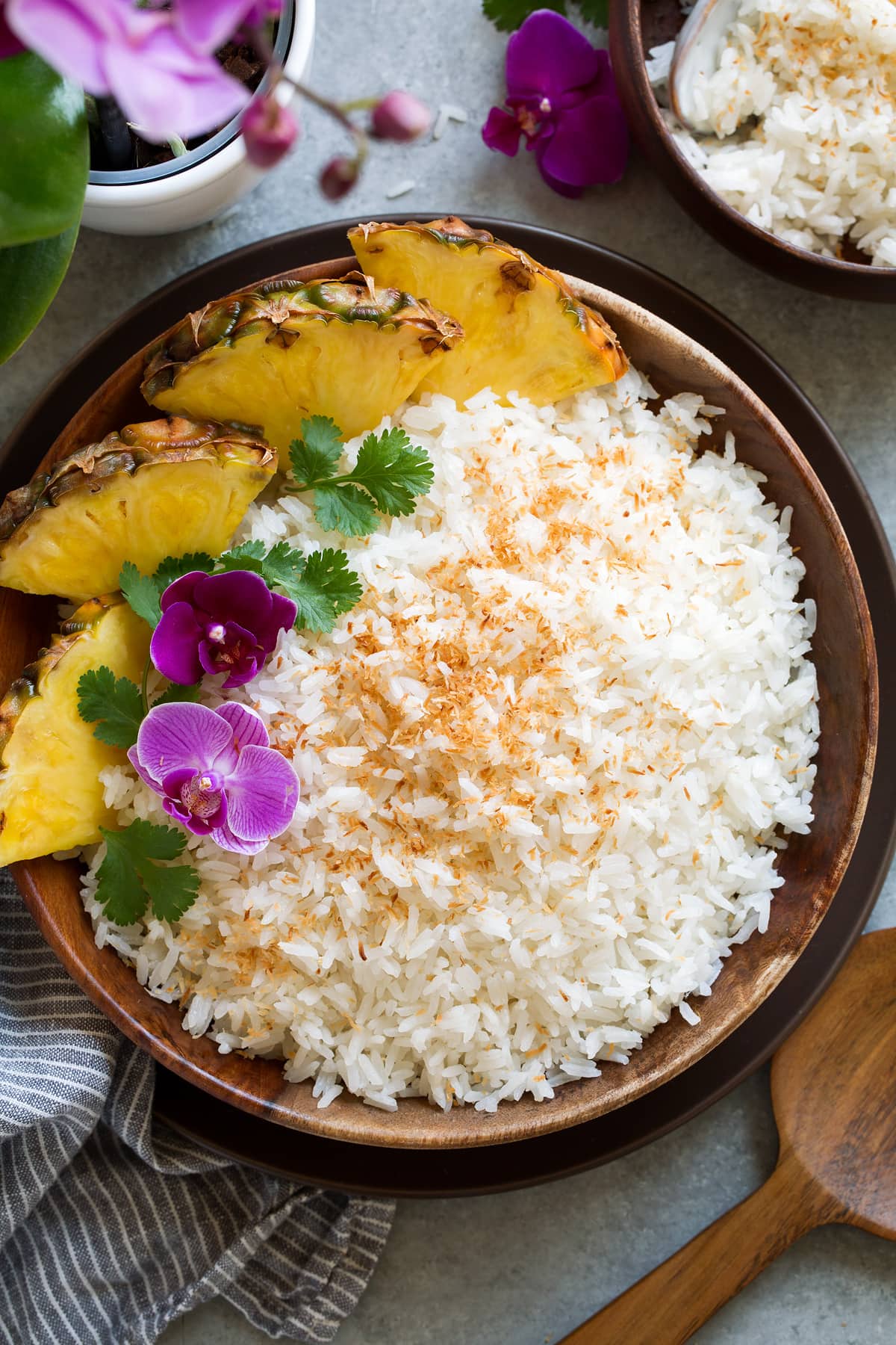 Wooden bowl full of Coconut Rice topped with flowers, cilantro and a side of fresh pineapple.