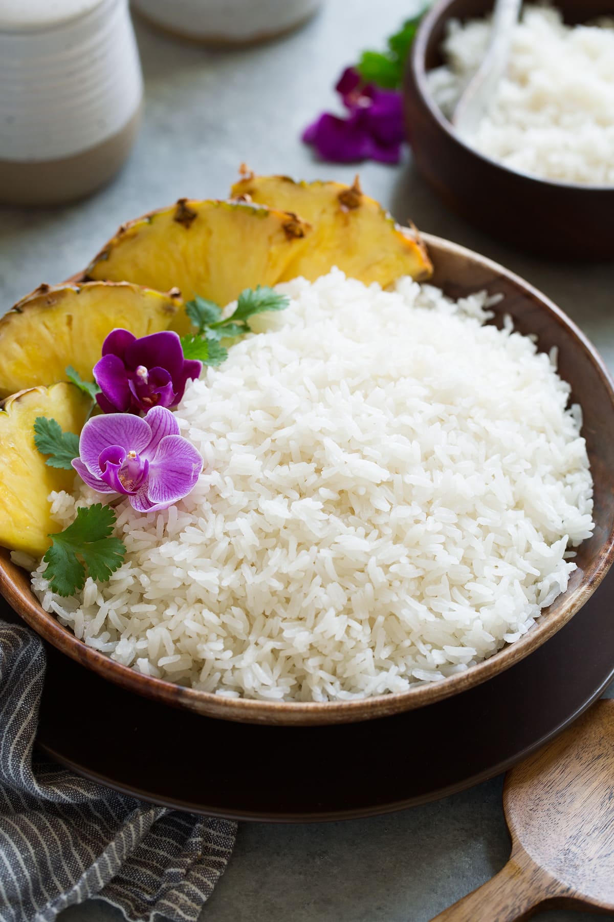 Coconut rice in a wooden serving bowl garnished with orchids, cilantro and pineapple wedges on the side.