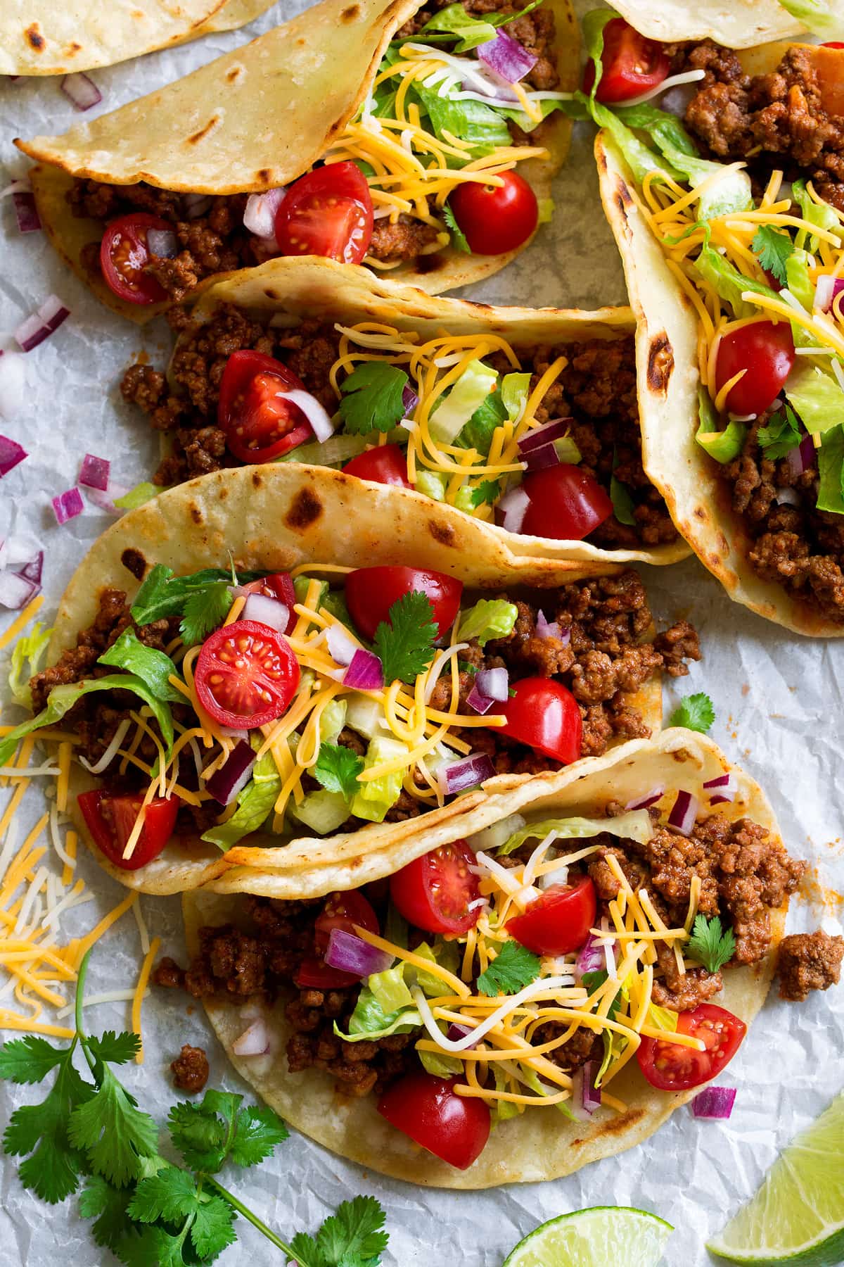 Row of tacos filled with ground beef filling, cheese, lettuce, tomatoes, and cilantro.