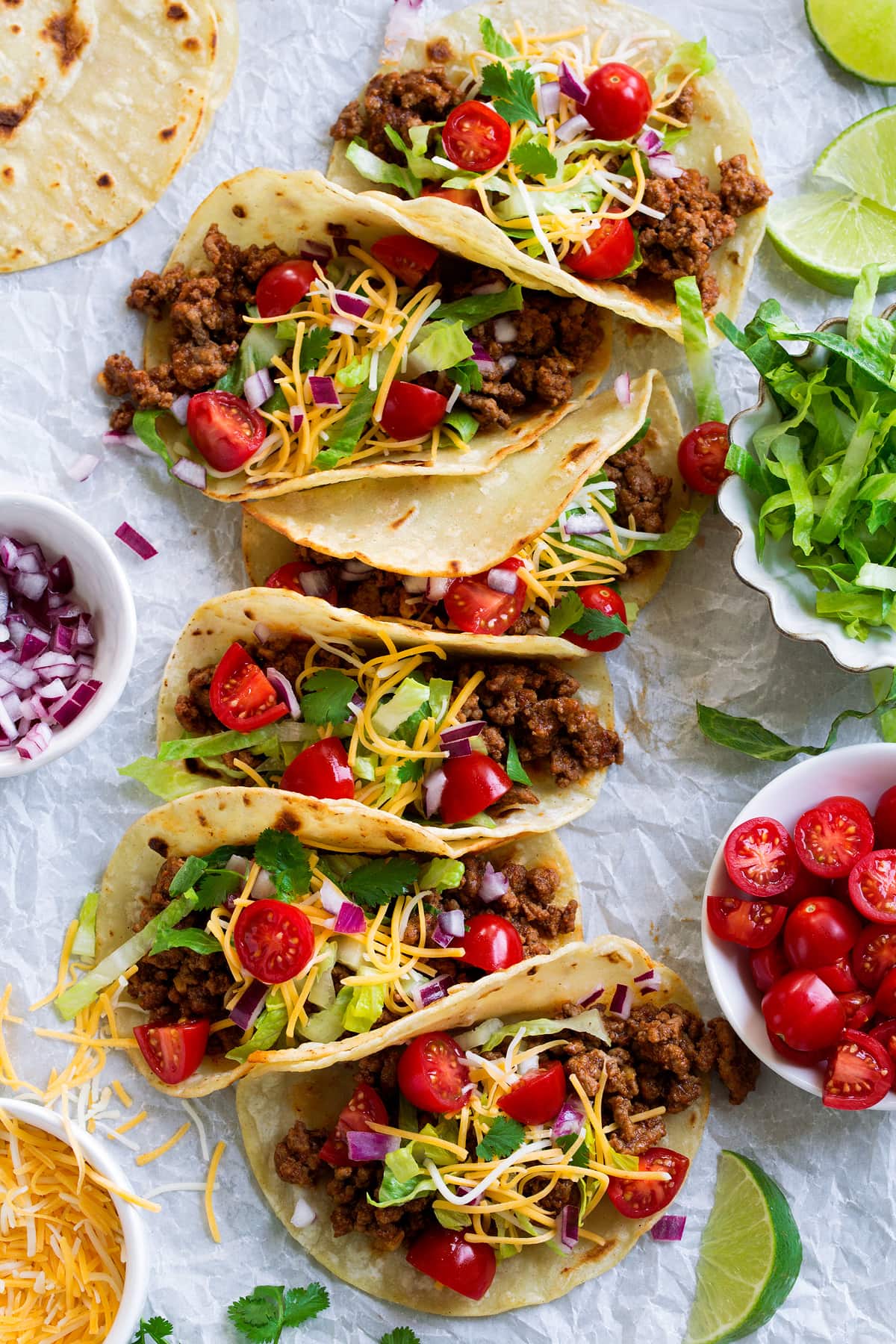 Long row of ground beef tacos on parchment paper finished with fresh toppings.