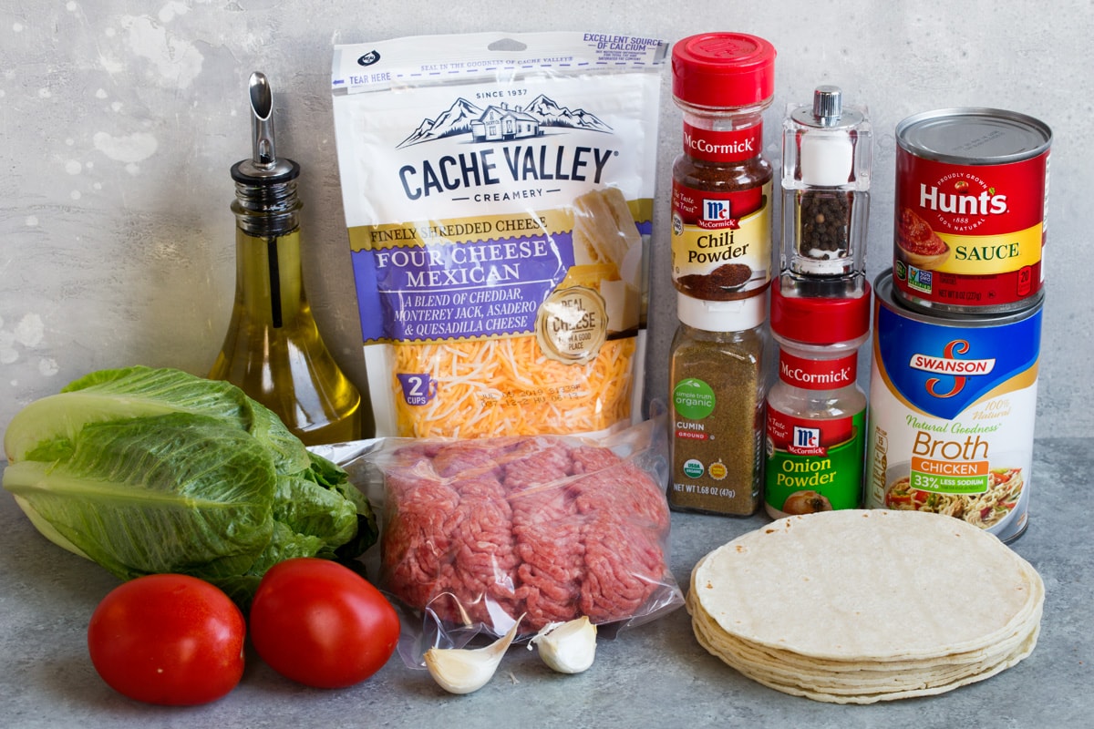 Ingredients needed to make tacos shown here including ground beef, oil, spices, tomato sauce, beef broth, tortillas, cheese, lettuce, garlic tomatoes.