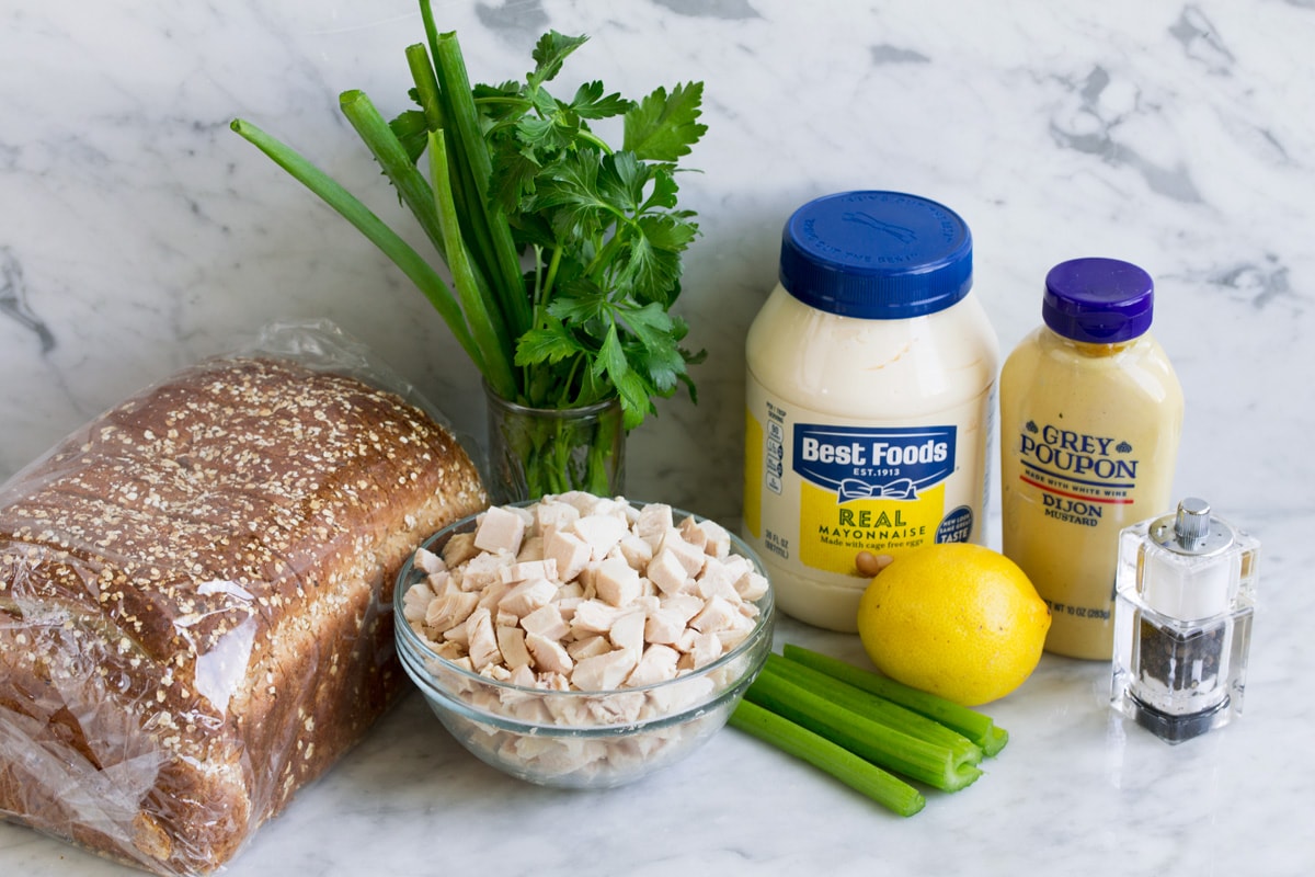 Chicken salad ingredients on a marble surface including cooked chicken breast pieces, celery, lemon, mayonnaise, dijon mustard, parsley, green onion, salt and pepper and bread for serving.