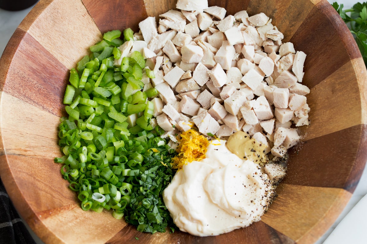 Showing how to make chicken salad, tossing chicken breast pieces, mayonnaise, dijon, lemon, celery, green onion, parsley in a wooden mixing bowl.