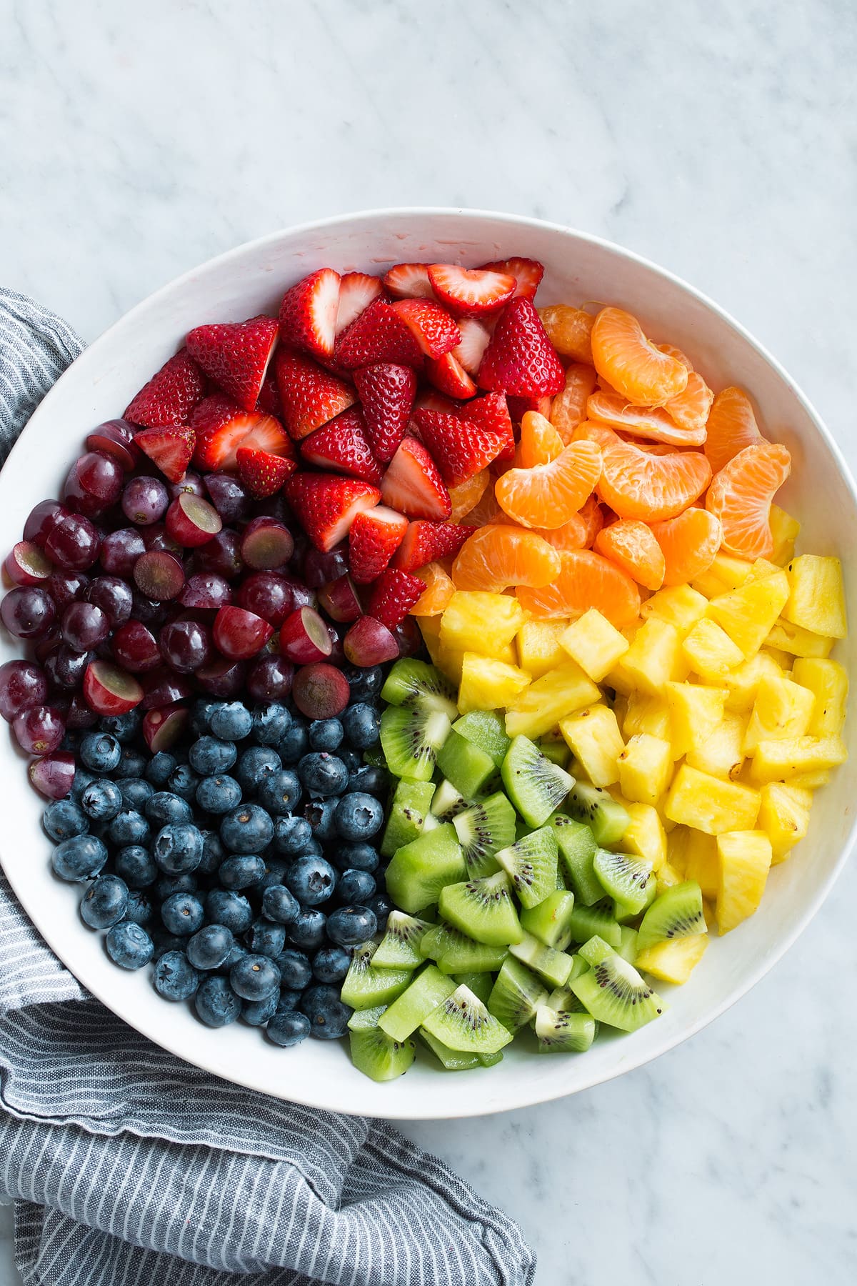 Fresh strawberries, mandarin oranges, pineapple, kiwi, blueberries, and grapes in sections in a large white serving bowl.