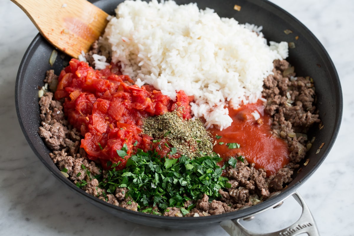 Adding cooked rice, tomatoes, tomato sauce, and herbs to ground beef mixture in skillet.