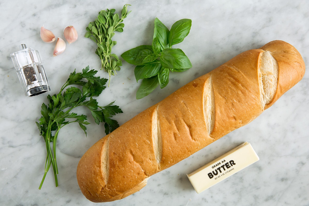 Ingredients for garlic bread including french bread loaf, garlic, butter, and fresh herbs.