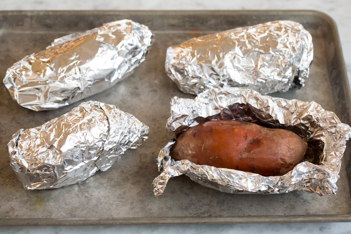 Four sweet potatoes wrapped in aluminum foil, one open, sitting on a baking sheet.