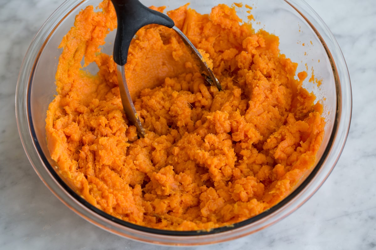 Mashing sweet potatoes in a glass mixing bowl with a potato masher. 