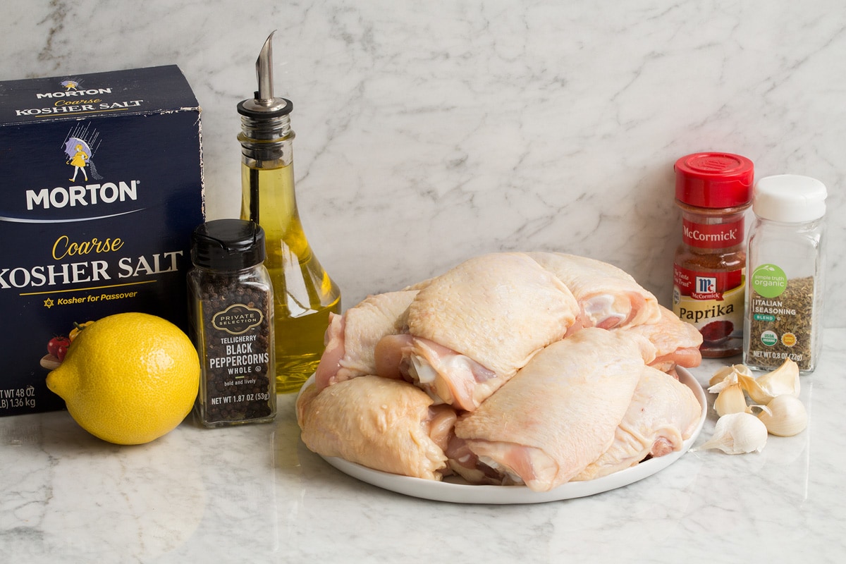 Image of ingredients needed to make baked chicken thighs. Shows raw chicken thighs, salt and pepper, olive oil, lemon, paprika, Italian seasoning and garlic.