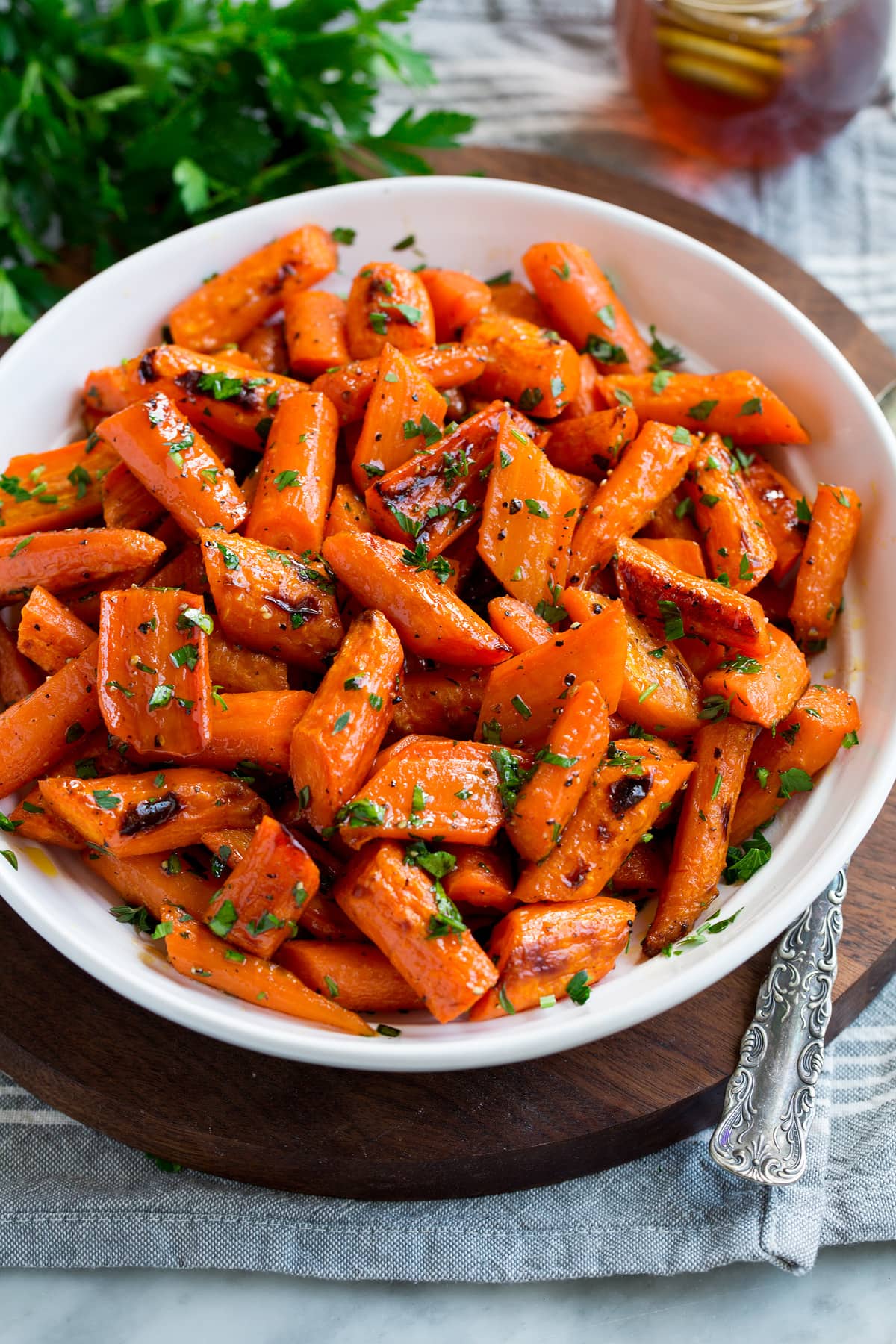 Photo: Roasted carrots in a white serving bowl. Carrots are garnished with parsley with a bunch in the background. Bowl is resting on a wooden platter and a grey cloth.