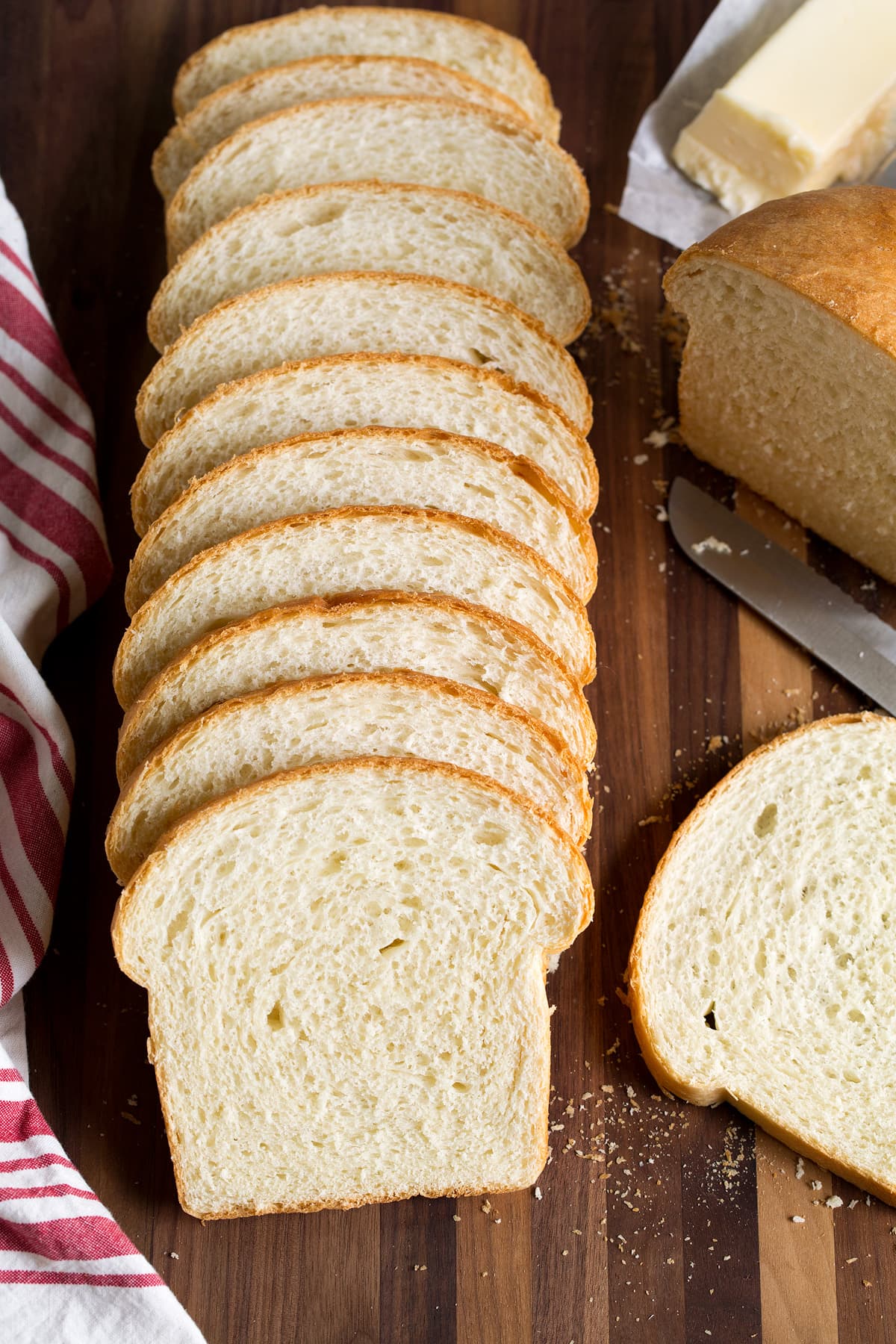 Row of fresh fluffy white bread on a cutting board with a kitchen cloth to one side and a knife and butter on the other side.