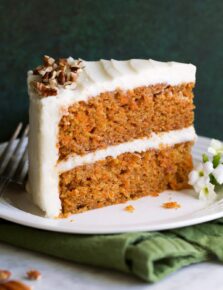 Slice of carrot cake with cream cheese frosting on a white dessert plate sitting on a green cloth napkin.