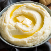 Mashed potatoes in a grey serving bowl over a grey napkin. Potatoes are topped with butter, shown as a serving suggestion.