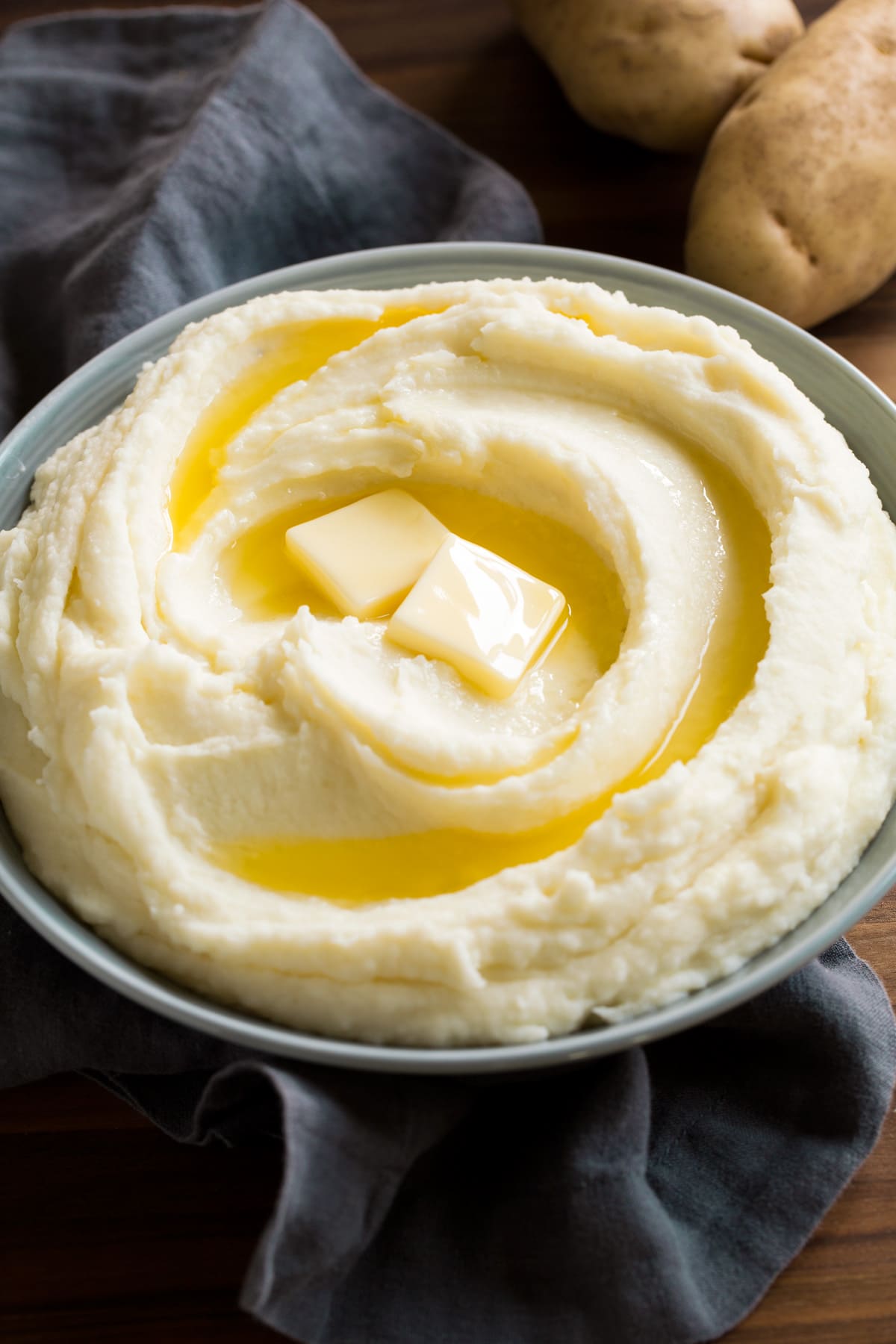 Mashed potatoes in a grey serving bowl over a grey napkin. Potatoes are topped with butter, shown as a serving suggestion.