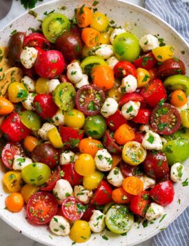 Tomato Salad in a white serving bowl. Shown in salad are multi-color cherry tomatoes and grape tomatoes, mozzarella pearls and it's coated in a herb dressing.