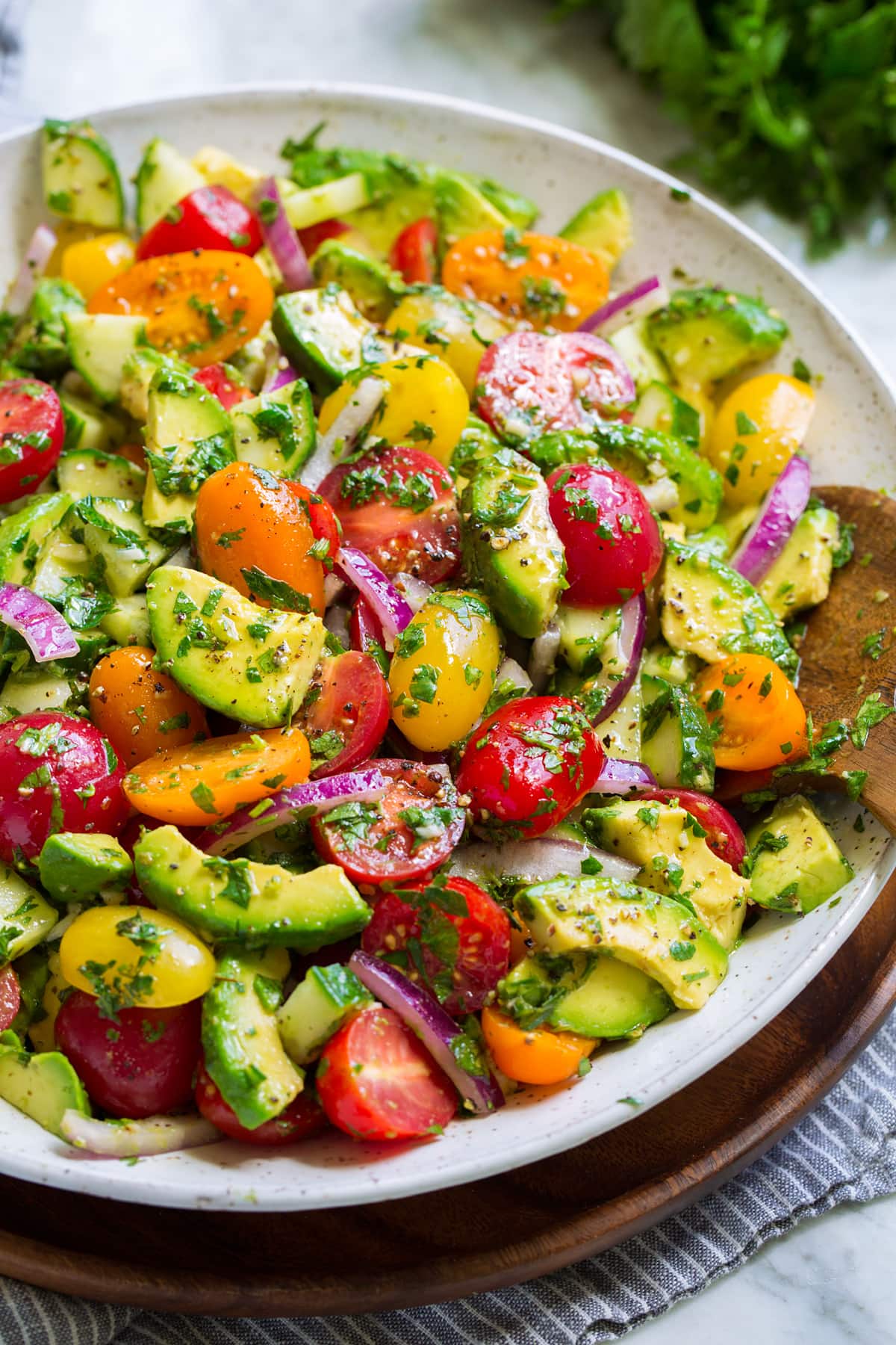 Avocado salad shown in a white serving bowl set over a wooden plate on a marble surface. Shown from a side angle.