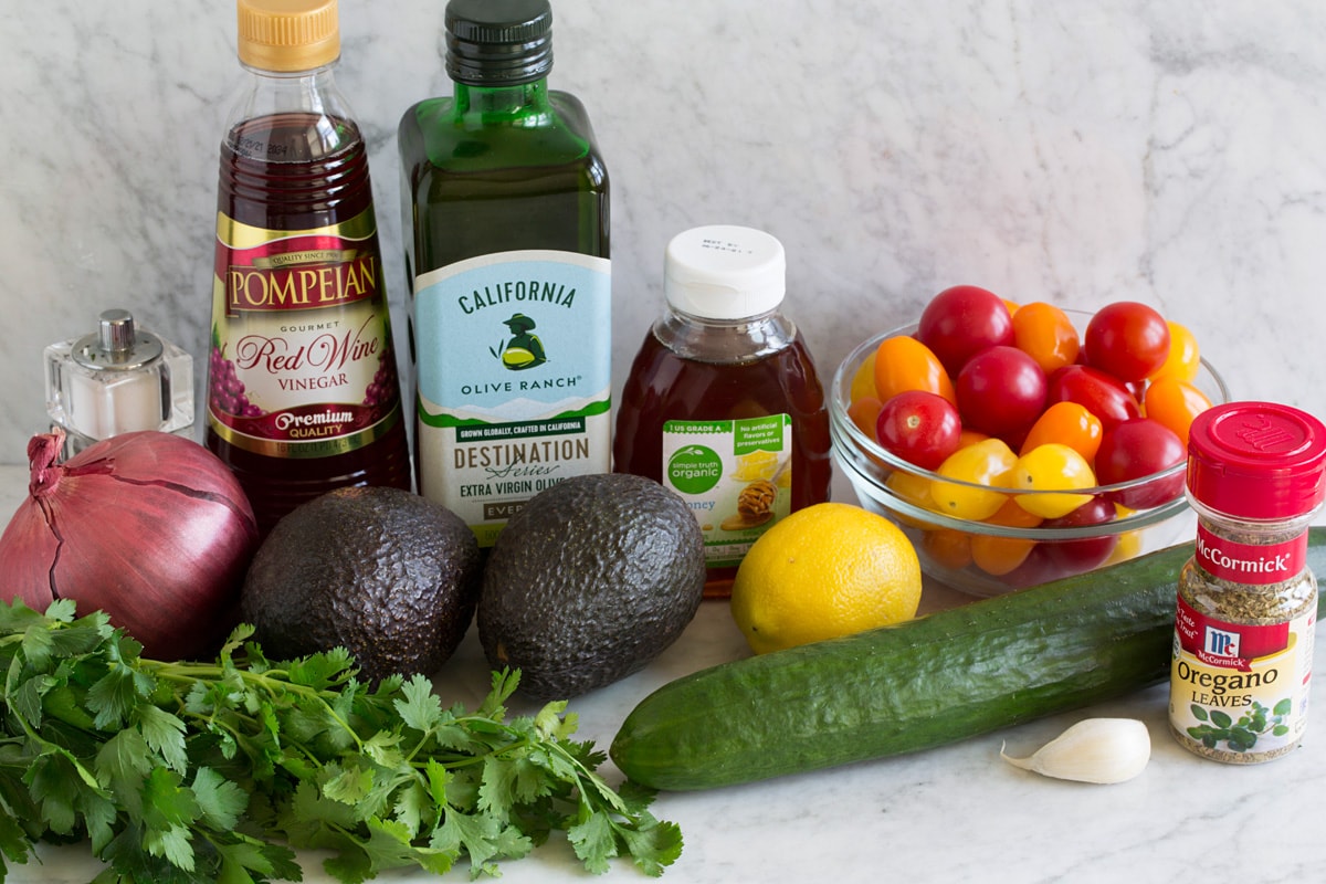 Image of ingredients that go into avocado salad. Includes fresh avocados, red onio, cilantro, parsley, cucumber, garlic, lemons, grape tomatoes, honey, olive oil, red wine vinegar and oregano.