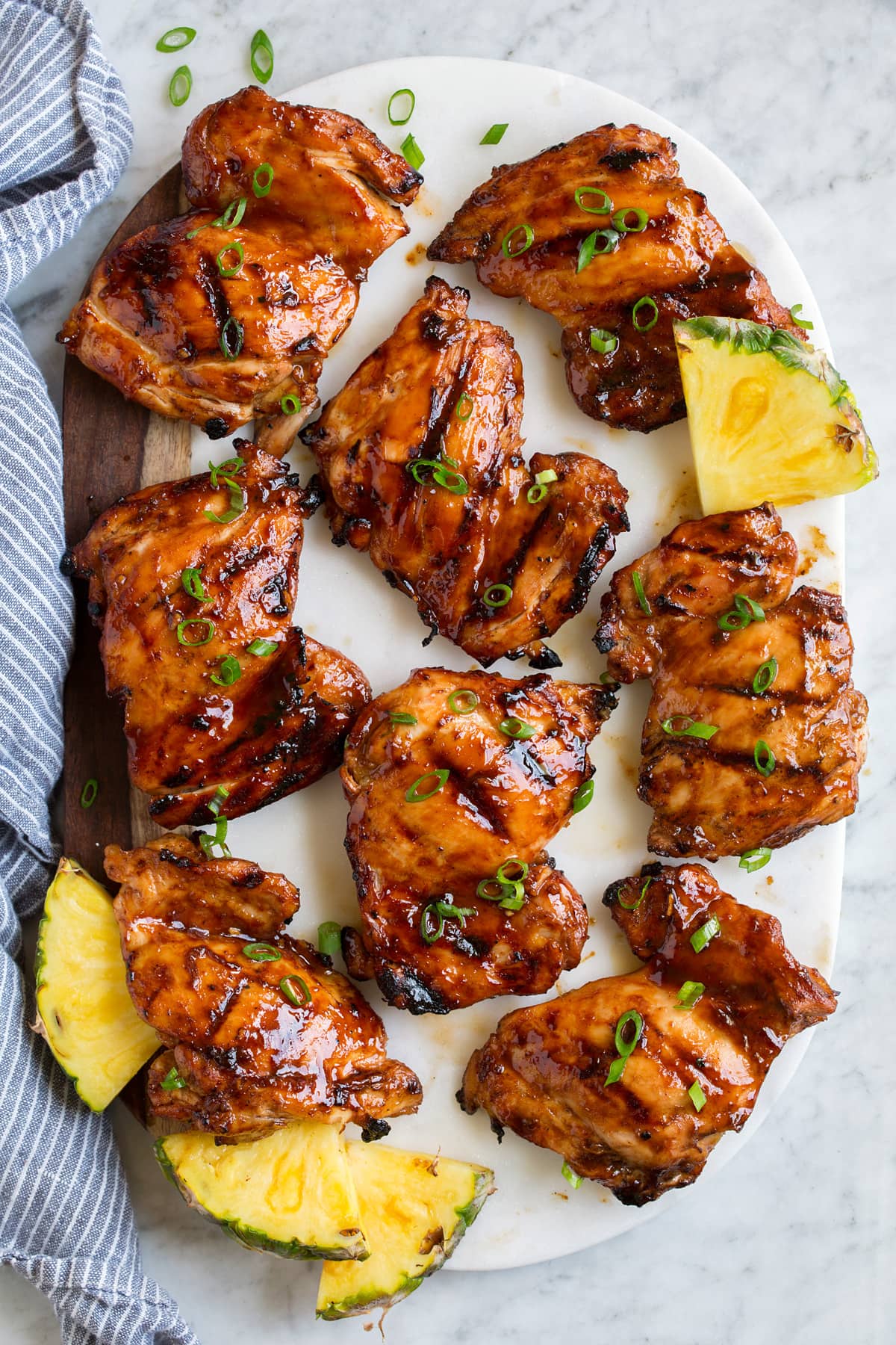 8 pieces of Huli Huli Chicken on a white marble and wood platter with pieces of pineapple on the side.