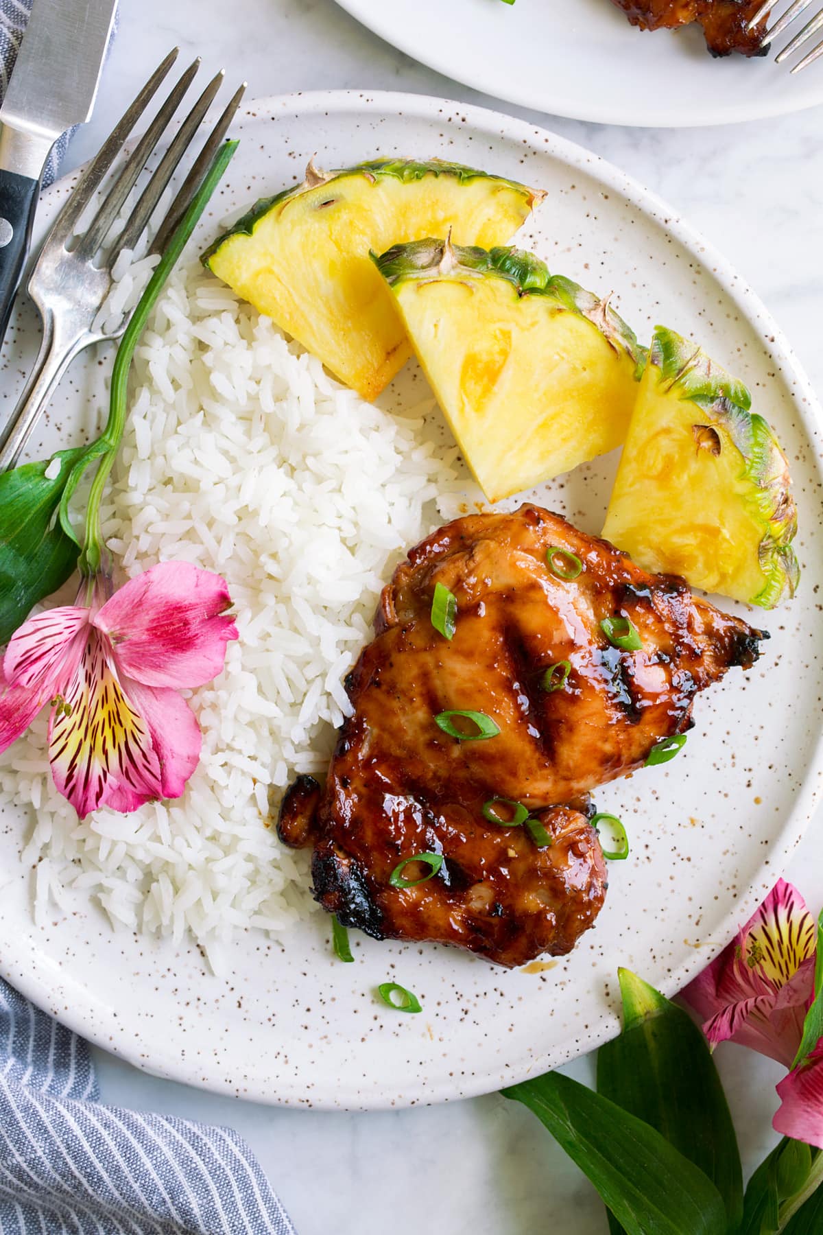 Plate with 1 huli huli chicken piece, pineapple, rice and a fresh flower. Shown overhead.