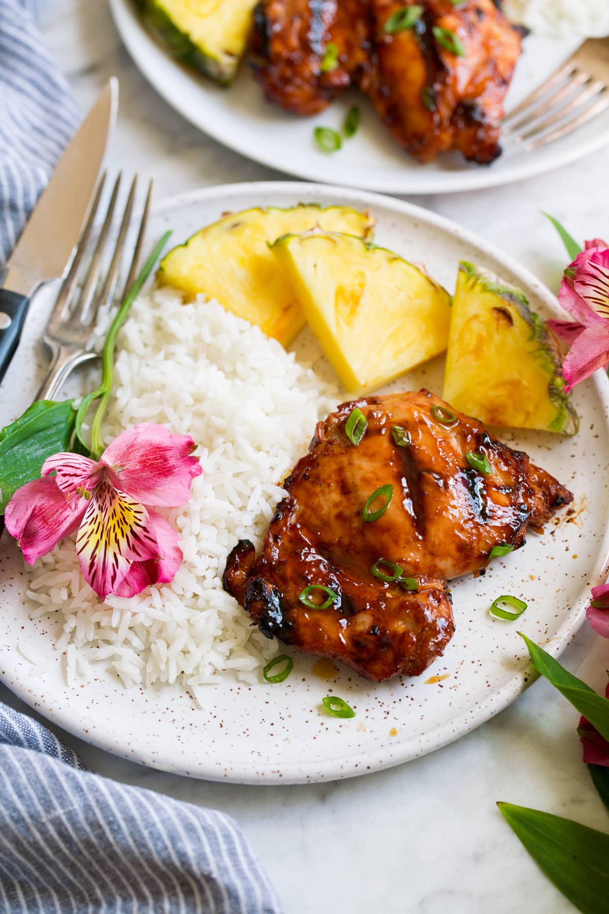 Plate with 1 huli huli chicken piece, pineapple, rice and a fresh flower. Shown from the side.