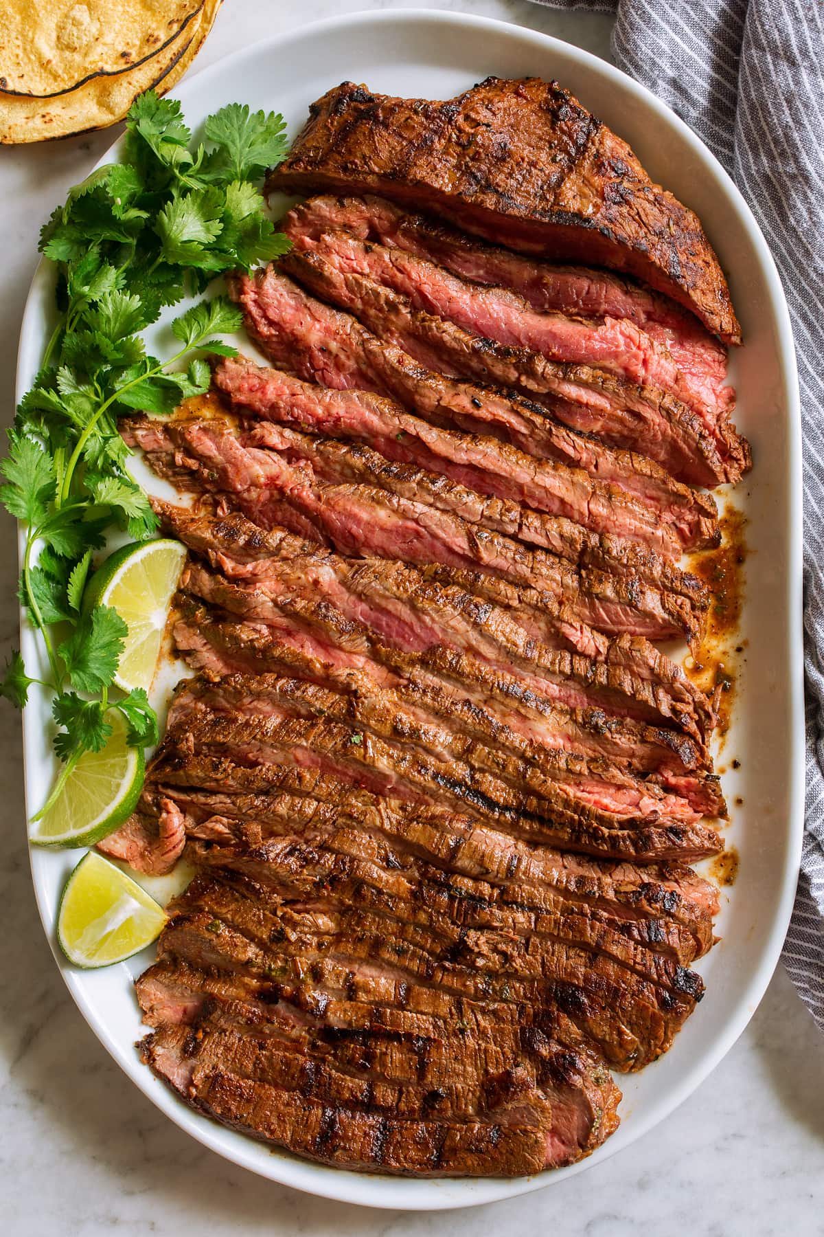 Overhead image of sliced carne asada on an oval white serving platter with cilantro, limes and tortillas on the side.