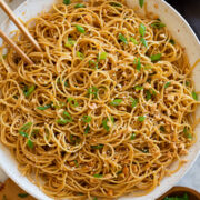 Photo: Bowl of sesame noodles topped with peanuts, sliced green onions and sesame seeds. Shown overhead in a white bowl with a dark napkin underneath and chopsticks and serving spoons to the side.