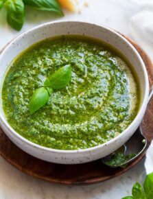 Bowl full of fresh homemade pesto. Bowl is resting on a wooden platter. Parmesan, fresh basil and pine nuts are shown in the background.