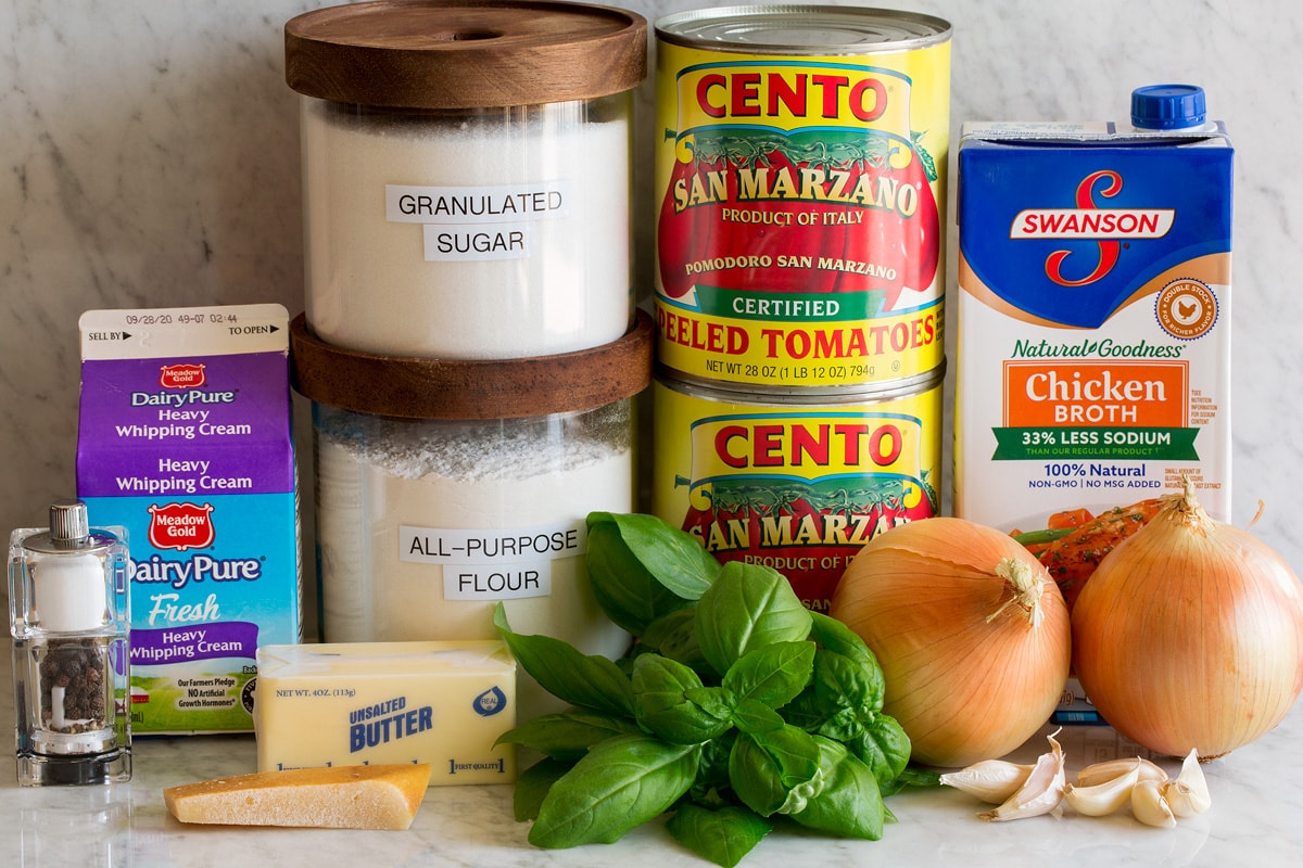 Image of ingredients used to make homemade tomato soup. Includes canned san marzano tomatoes, chicken broth, onions, garlic, basil, flour, sugar, butter, parmesan rind, cream, salt and pepper.