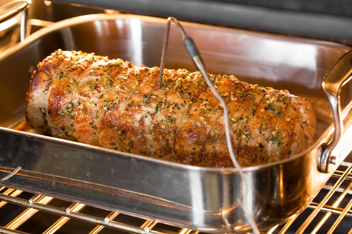 Pork loin shown in roasting pan in the oven with an oven probe thermometer inserted into center.