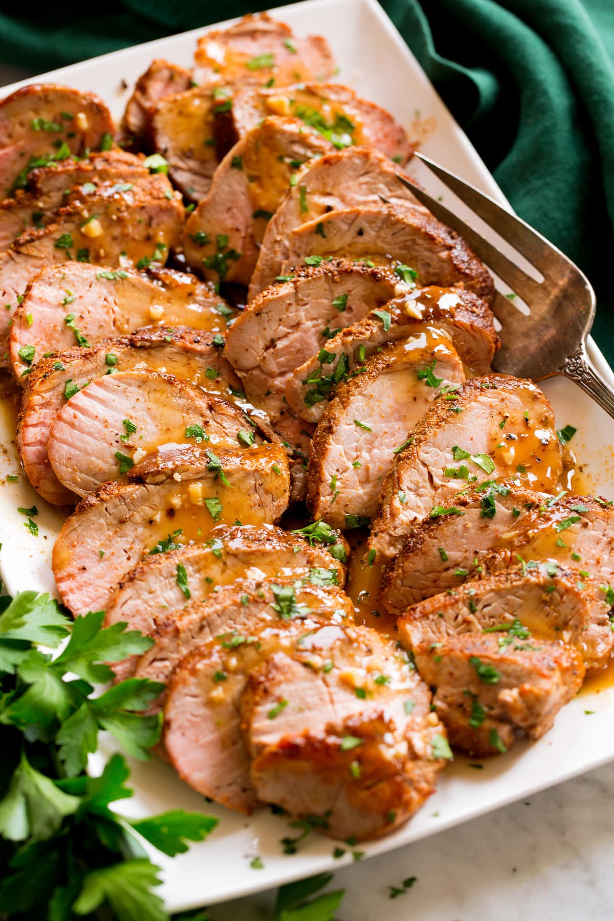 Baked Pork Tenderloin sliced into strips resting shown on a platter. Pork is rubbed with spices, topped with a pan sauce and parsley.